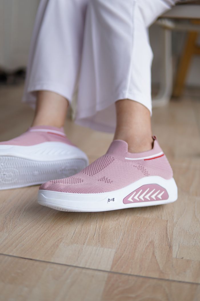 Trendy pink slip-on sneakers worn by a woman with white pants on wooden floor.